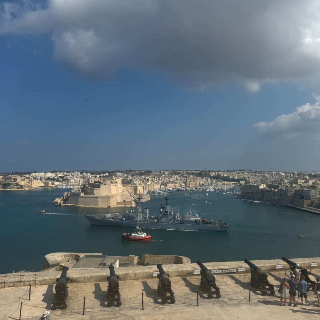 Saluting battery in Malta, cannons lined up in front of Valletta's Grand Harbour. An Italian navy ship is sailing by. Four people are standing behind the cannons looking at them in the lower right-hand corner.