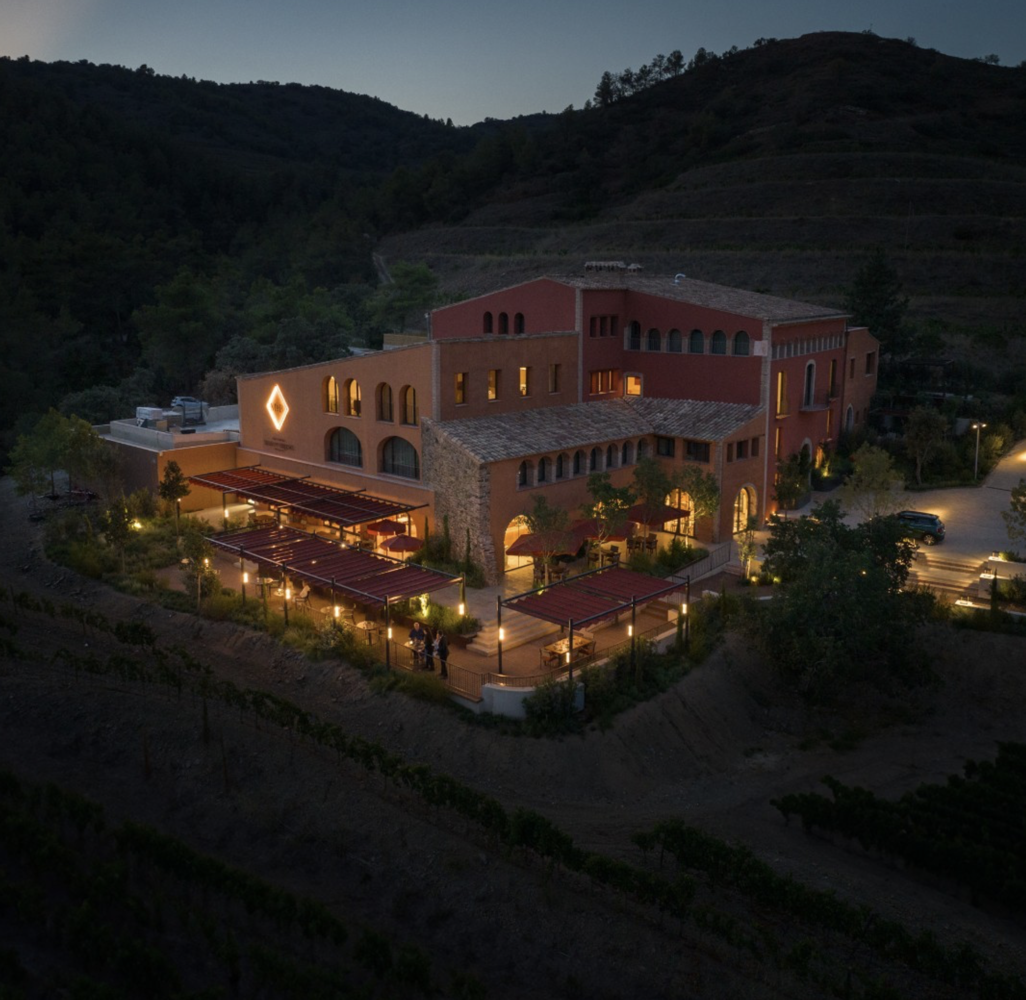 Night time view of Gran Hotel Mas d'en Bruno, can see the building with the lights on giving it a golden glow. It's set in the foreground, with some hills behind it. Night time.