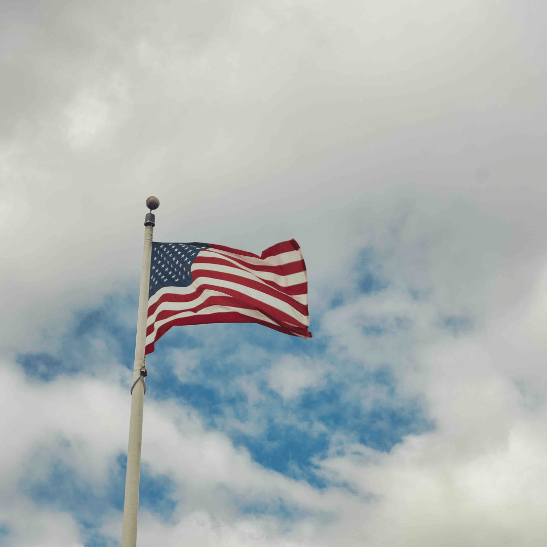 United States flag waving in the wind, can see a blue sky and clouds behind it. Camera is facing upwards.