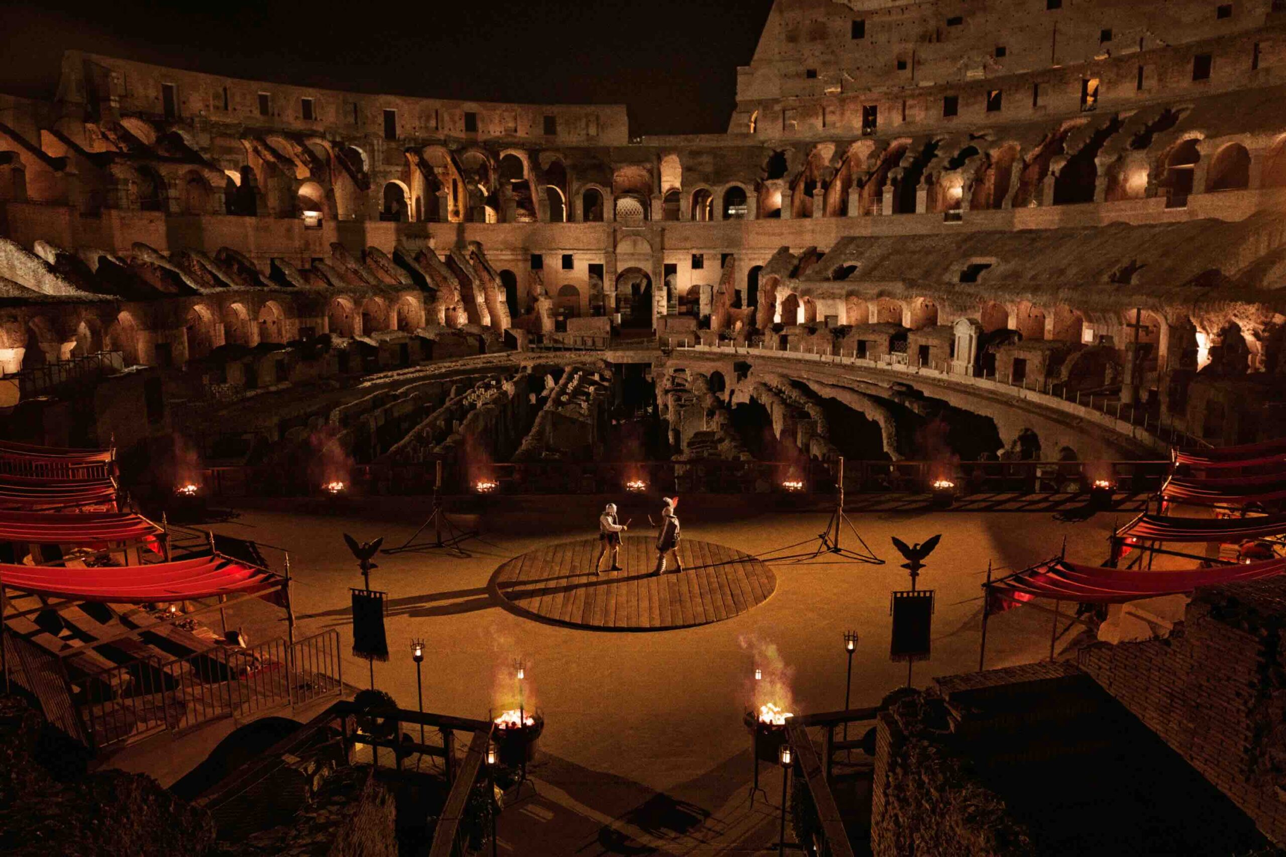 The inside of the Colosseum in Rome. Can see candles lit around the outside and two Gladiators in the center.